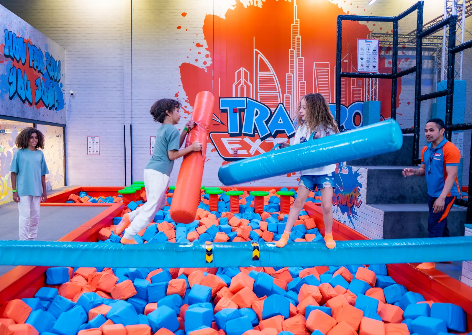 Two challengers battling with jousting sticks on the beam over a foam pit at Trampo